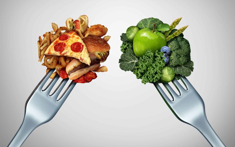 42846553 - diet struggle and decision concept and nutrition choices dilemma between healthy good fresh fruit and vegetables or greasy cholesterol rich fast food with two dinner forks competing to decide what to eat.